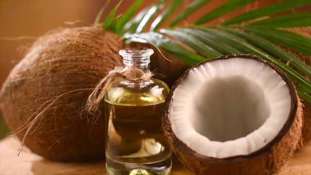 Coconut oil in a bottle with coconuts and green palm leaf rotated over brown background. Healthy food, skincare concept. Rotation 4K UHD video 3840X2160