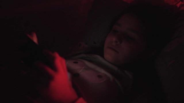 Little girl lying in the dark under a blanket looking at smartphone. Red light in the room