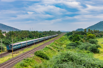 awesome view of indian railway running on track goes to horizon in green landscape under blue sky with clouds.