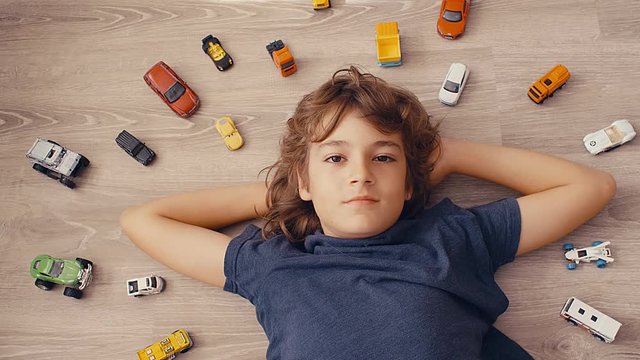 Top view of a 10-years boy lying on the ground and dreaming with toy cars around, 4k slow motion