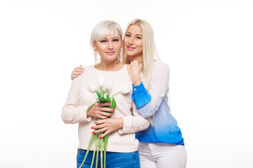 Teen blond daughter and mature mother with bouquet of flowers on white background hugging