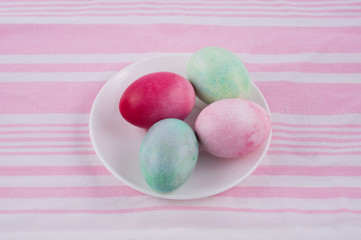 four multicolored painted Easter eggs on a white plate and  striped pink-white tablecloth. holiday easter greeting card, poster, wallpaper
