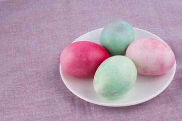 four multicolored painted Easter eggs on a white plate and lilac tablecloth. holiday easter greeting card, poster, wallpaper