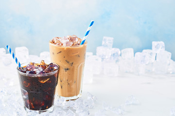 Summer drink iced coffee in a glass and ice coffee with cream in a tall glass surrounded by ice on white marble table over blue background. Selective focus, copy space for text. Horizontal.