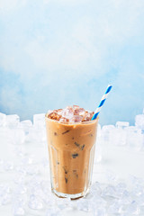 Summer drink ice coffee with cream in a tall glass with straw surrounded by ice on white marble...