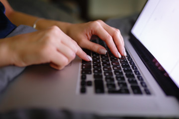 Close-up of woman's hand using laptop with blank screen on bed in home interior. The light from the screen illuminates the female hands on the laptop keyboard. Freelancer works at home in the evening