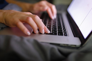 Close-up of woman's hand using laptop with blank screen on bed in home interior. The light from the screen illuminates the female hands on the laptop keyboard. Freelancer works at home in the evening