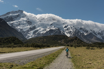 Fototapeta na wymiar Female, baby boomer cyclist cycling on a bike path towards the snow capped and glacier covered Southern Alps, Aoraki/Mount Cook National Park, New Zealand.