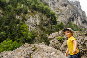Naklejka na ściany i meble Lateral view of a 6-years blond boy near a rocky mountain surrounded by grass