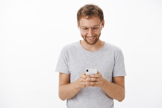 Texting with girlfriend being on way home. Relaxed and joyful handsome aduly male with beard in glasses and gray t-shirt looking down at smartphone screen with broad smile having fun messaging
