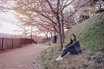 Naklejka na ściany i meble Happy young asian woman sitting on green grass in park under cherry blossom tree relaxing in Japan. Relax and lifestyle concept.