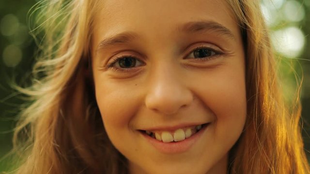 Close up of a pretty small blonde girl's face smiling straight into the camera on the blurred park background. Portrait. Outside