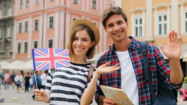 Travelling young couple using local map on a sunny day with the landmarks of the town in the background. Young pretty woman holding british flag and waving it happily. Outdoors.