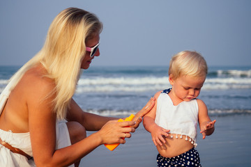 mother in stylish glasses and white dress is putting orange spray bottle SPF on newborn baby one...
