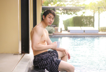 Fototapeta na wymiar asian man sitting on the edge of a swimming pool and holding cup of coffee