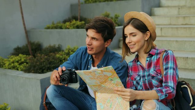 Portrait of a handsome boy and a young pretty woman on vacation trip. Caucasian couple holding a map and a camera while sitting on stairs outdoor.