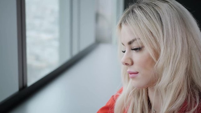 Portrait of Woman in red coral business suit. Young blonde businesswoman with modern gray laptop in the office in high floor in front of the window. Girl drinking coffee