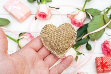 Hand holding decorative burlap heart over table with roses and gift box. Valentine day card.