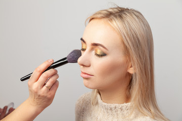 Makeup artist puts make-up on a blonde model with eyes closed, overlay the shadows in the Oriental styleholds a brush in her hands and applies blush to her face on a white background in visage studio.