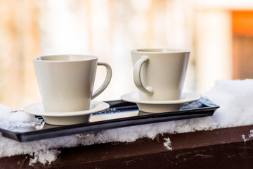 Two beige mugs with hot drinks, on the edge of a balcony, with snow on the sides and a snowy nature in the background.Winter still life with cup of hot drink and snowy fir tree background
