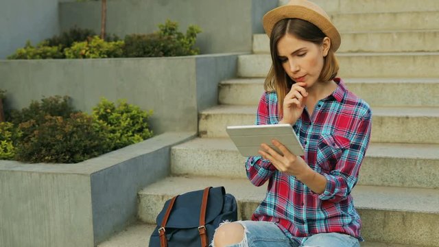 Young stylish girl sits on steps and holding hands jn the tablet. Girl student communicates in social networks with friends by typing messages with stairs in the background.