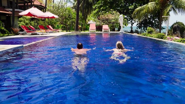 man and woman swims in swimmingpool in luxury hotel. couple leisure in the pool luxury hotel with swimming pool, sun beds, palm trees by sea. tropical resort with pool. Travel concept.