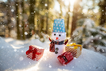 Happy snowman with gift boxes standing in winter christmas landscape. Snowman smiling standing in snow near spruce trees