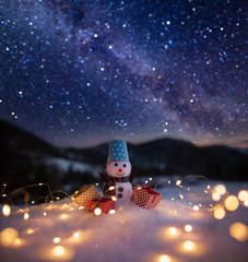 Happy snowman with gift boxes standing in winter christmas landscape. Snowman in mountains. Space landscape with silhouette mountains. Milky Way at mountains