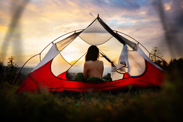 Naked woman sitting in tent in sleeping bag and enjoy the scenery in the mountains. Incredible cloudy sky at sunrise or sunset. Trekking concept. Relaxing, feeling alive, got freedom from work