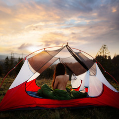 Naked woman sitting in tent in sleeping bag and enjoy the scenery in the mountains. Incredible cloudy sky at sunrise or sunset. Trekking concept. Relaxing, feeling alive, got freedom from work