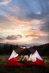 Naked woman sitting in tent in sleeping bag  and drinking coffee in the morning. A young female tourist drinks a hot drink and enjoy the scenery in the mountains at sunrise or sunset