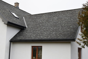 Part of white house with new wooden eaves, plastic rain gutter, window and chimney in grey shingles roof and brown window in facade.