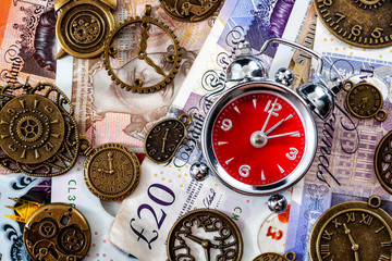 Time is money concept with flat lay of alarm clock on different denomination english pound bills or notes and surrounded by metal clocks