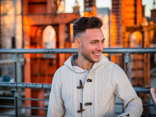 Handsome trendy young man smiling and laughing, standing on a sidewalk wearing a fashionable jacket and scarf