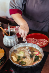 Woman stirring Hot and Boiling Shabu broth with cabbage, eryngii, enotitake, tofu and Kurobuta pork inside hot pot.
