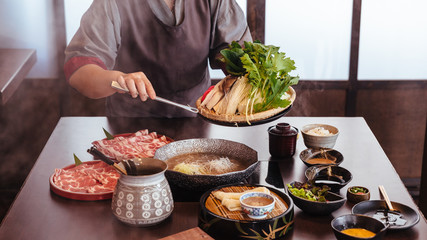 A woman pinching vegetables into hot pot by tongs with Wagyu A5 beef and sliced Kurobuta in Shabu clear soup with stream.