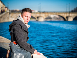 Fototapeta premium One handsome young man in urban setting in European city, Turin in Italy sitting on the banks of the river Po, in cold winter day