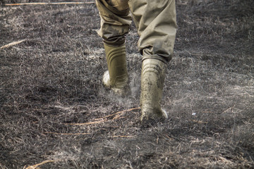 Legs in boots walking on burnt withered grass with ashes in the air, environmental problem of fires