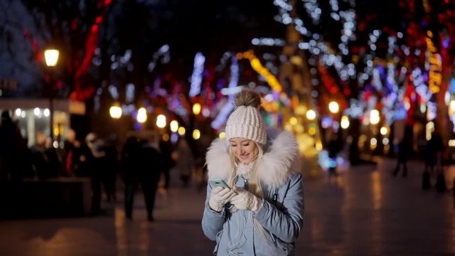 Cute and shy attractive young girl teenager uses her smartphone to listen to music and chat or connect with her friends, while relaxing in city on chilly winter day, Christmas bright lights background