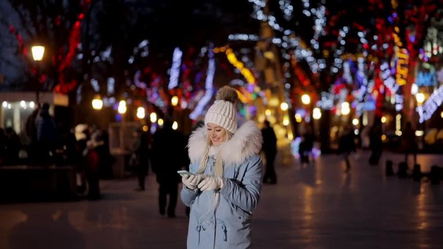 Portrait of young attractive girl in urban winter background listening to music with smartphone, New Year holidays, video in slow motion