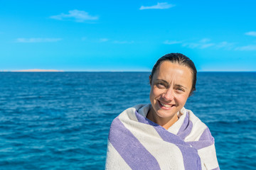 Fototapeta premium beautiful woman in a towel by the sea. Portrait of preteen brunette woman, wet and wrapped in towel after swimming, against blue sea and sky.