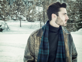 Stylish man walking confidently looking away on territory of contemporary winter resort covered in snow in the mountain, wearing brown leather jacket