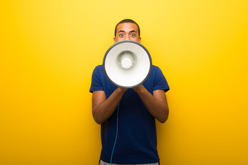 African american man with blue t-shirt on yellow background shouting through a megaphone to...