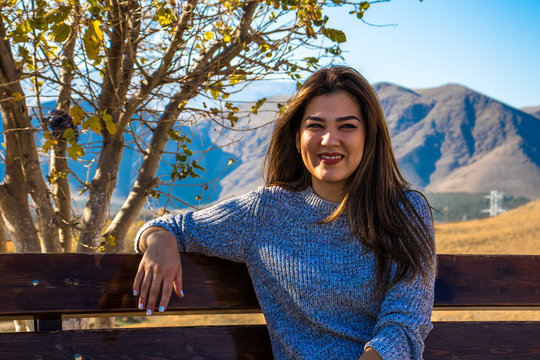 a young woman sitting on a bench and smiling on a background a tree covered with yellow leaves and a beautiful view on Kopet Dag mountains in Turkmenistan  