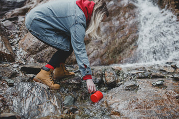 Beautiful girl traveler with mug is drawing water in waterfall