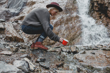 Beautiful girl traveler with mug is drawing water in waterfall