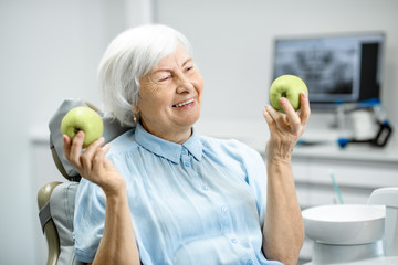 Portrait of a beautiful senior woman with healthy smile holding green apples at the dental office