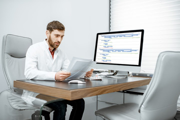 Obraz premium Handsome doctor reading some documents sitting in the luxury medical office with computer on the background