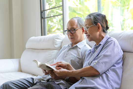 Asian senior couple sitting on sofa and reading a book in living room. They read the book with smile while wife pointing at the book and have some conversation . Happy retirement life concept.