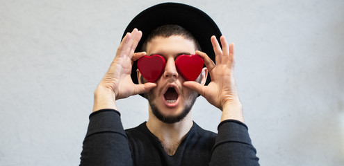 Shocked man holding red hearts on his eyes. Dressed in black, with hat. Valentines day background.